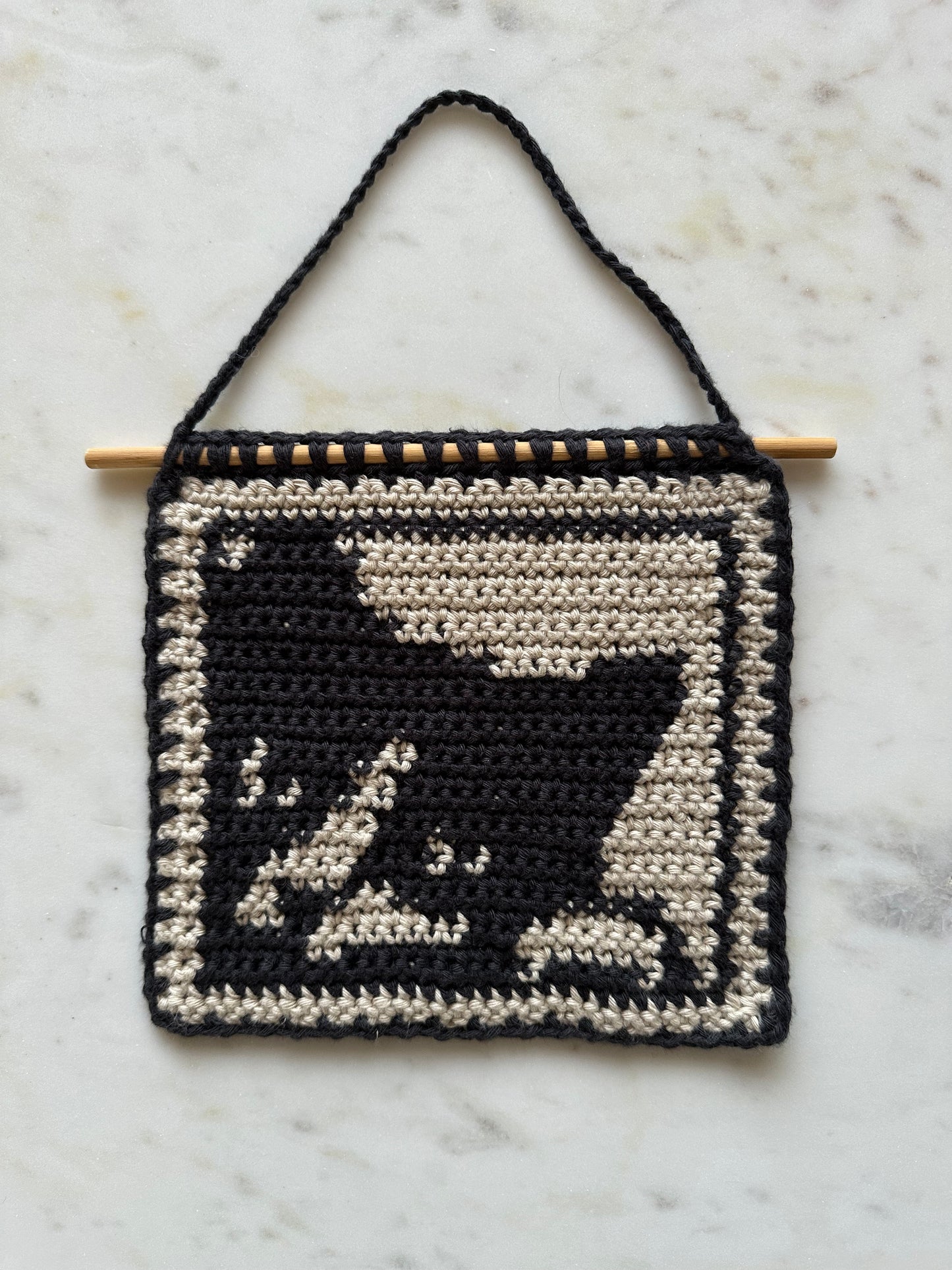 Shows our crocheted tapestry of a brown and black cat peeking through a frame. Made by The Woollers