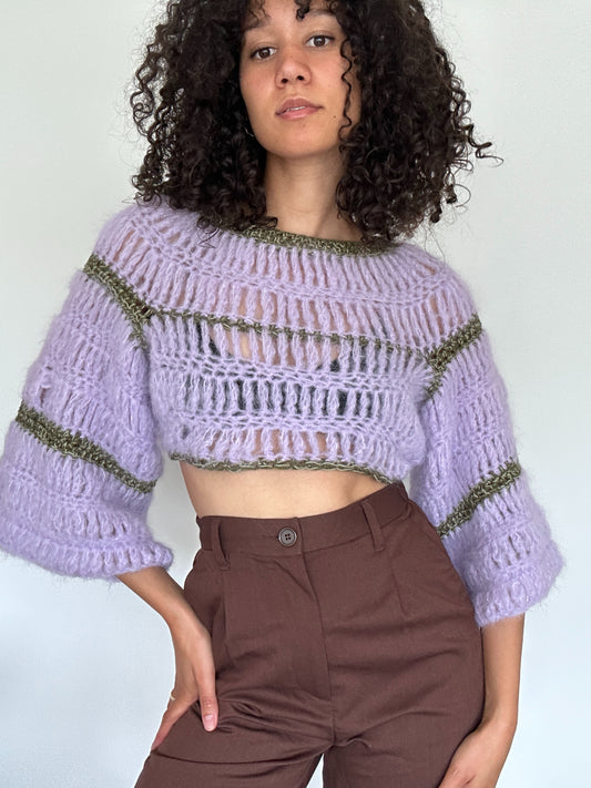Shows a model wearing a lila and green coloured FLORA Long Sleeve Crop Top, which is crocheted and has a flowy design. Made by The Woollers