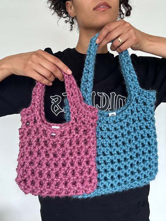 Shows a model holding two crocheted KNOTTY Handbags with an open mesh design, a smaller one in pink and a medium sized one in blue.  Made by The Woollers