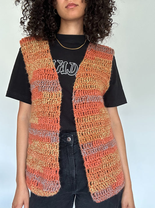Shows a model wearing the crocheted MARI Open Vest which has hues of orange, yellow and blue. Made by The Woollers