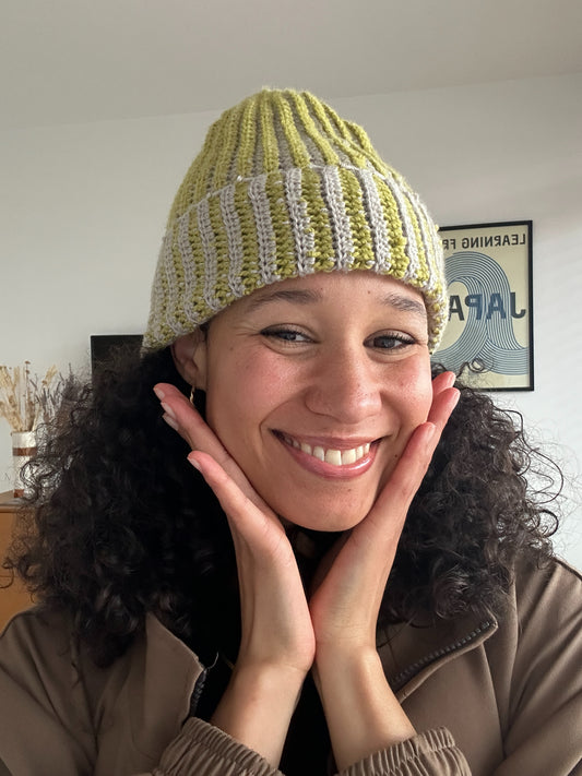 Shows a model wearing the reversible beanie while smiling. The beanie is striped with grey and green. Made by The Woollers