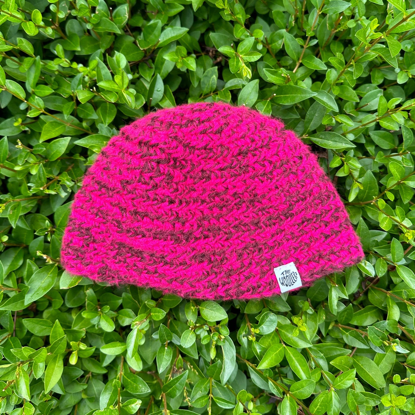 Shows a crocheted DOBI Beanie in the colors pink and purple with a label that says 'The Woollers'