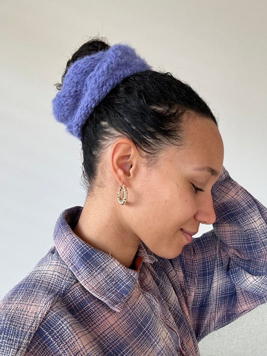 Shows a model from side view wearing the knitted MIMI Scrunchie XL. The scrunchie is knitted with a fluffy lila yarn. Made by The Woollers