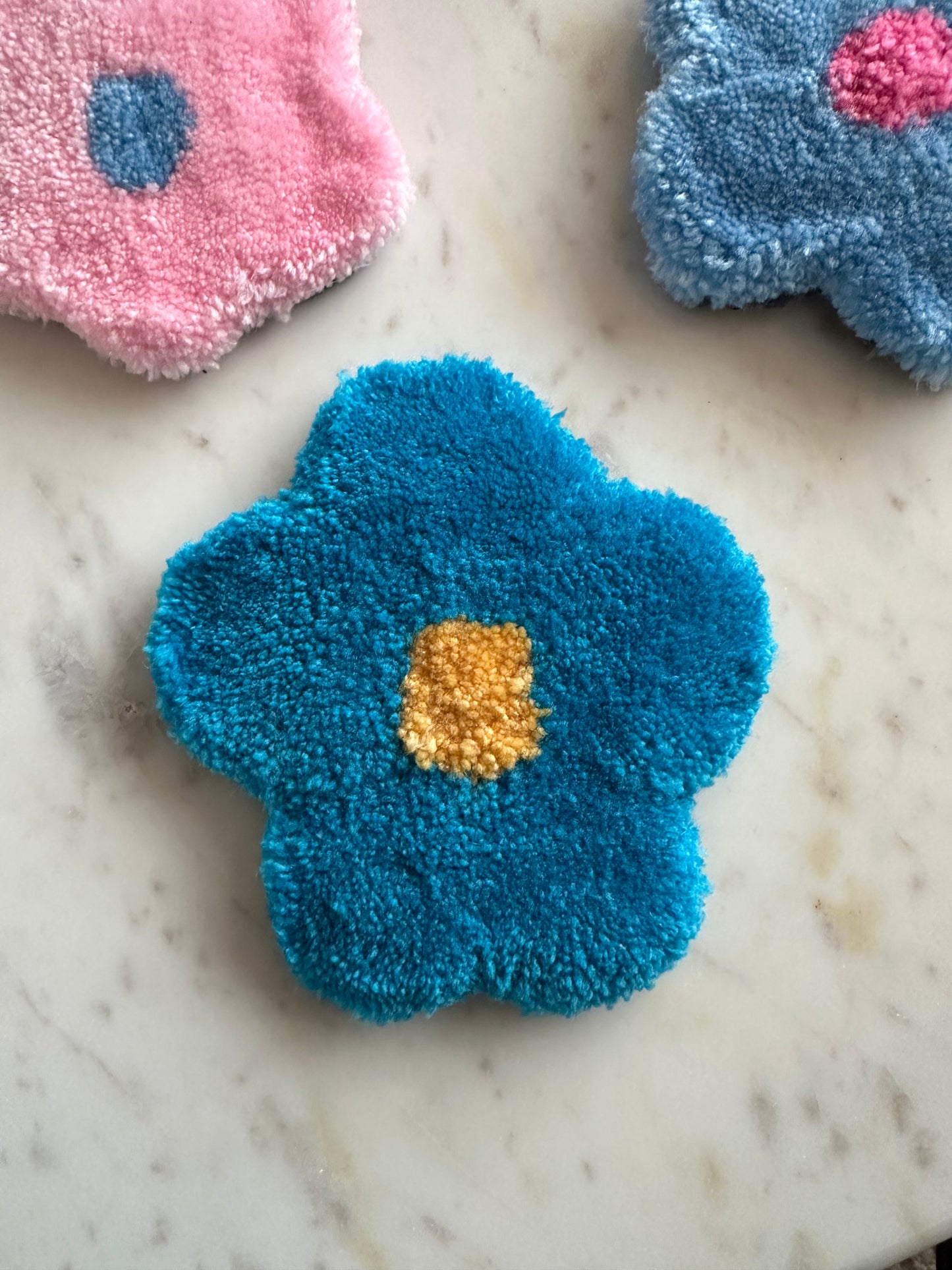 Shows one of our tufted flower coasters with blue petals and a yellow core. Made by The Woollers