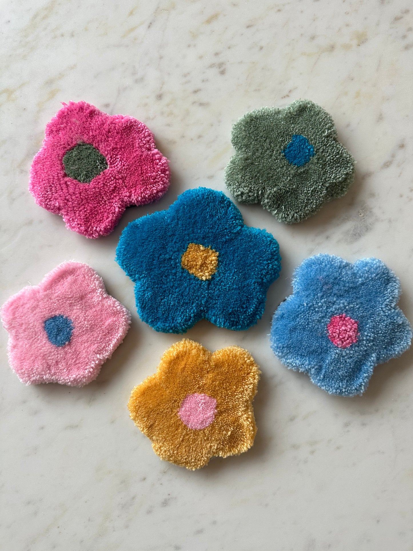 Our tufted flower shaped coasters placed on a table in various color combinations, such as green and blue, pink and green, yellow and pink, pink and blue, blue and pink. Made by The Woollers
