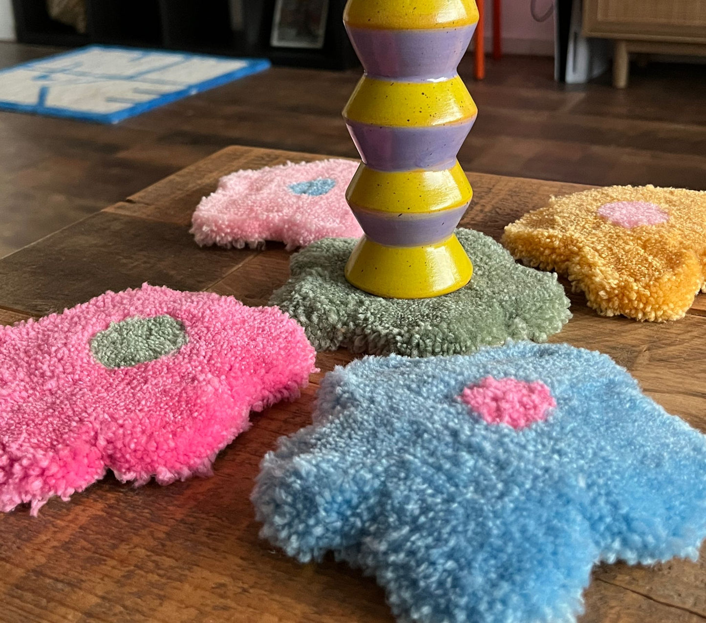 Our tufted flower shaped coasters placed on a table in various color combinations, such as green and blue, pink and green, yellow and pink, pink and blue, blue and pink. A yellow and lila candle holder is placed on one of the coastersMade by The Woollers