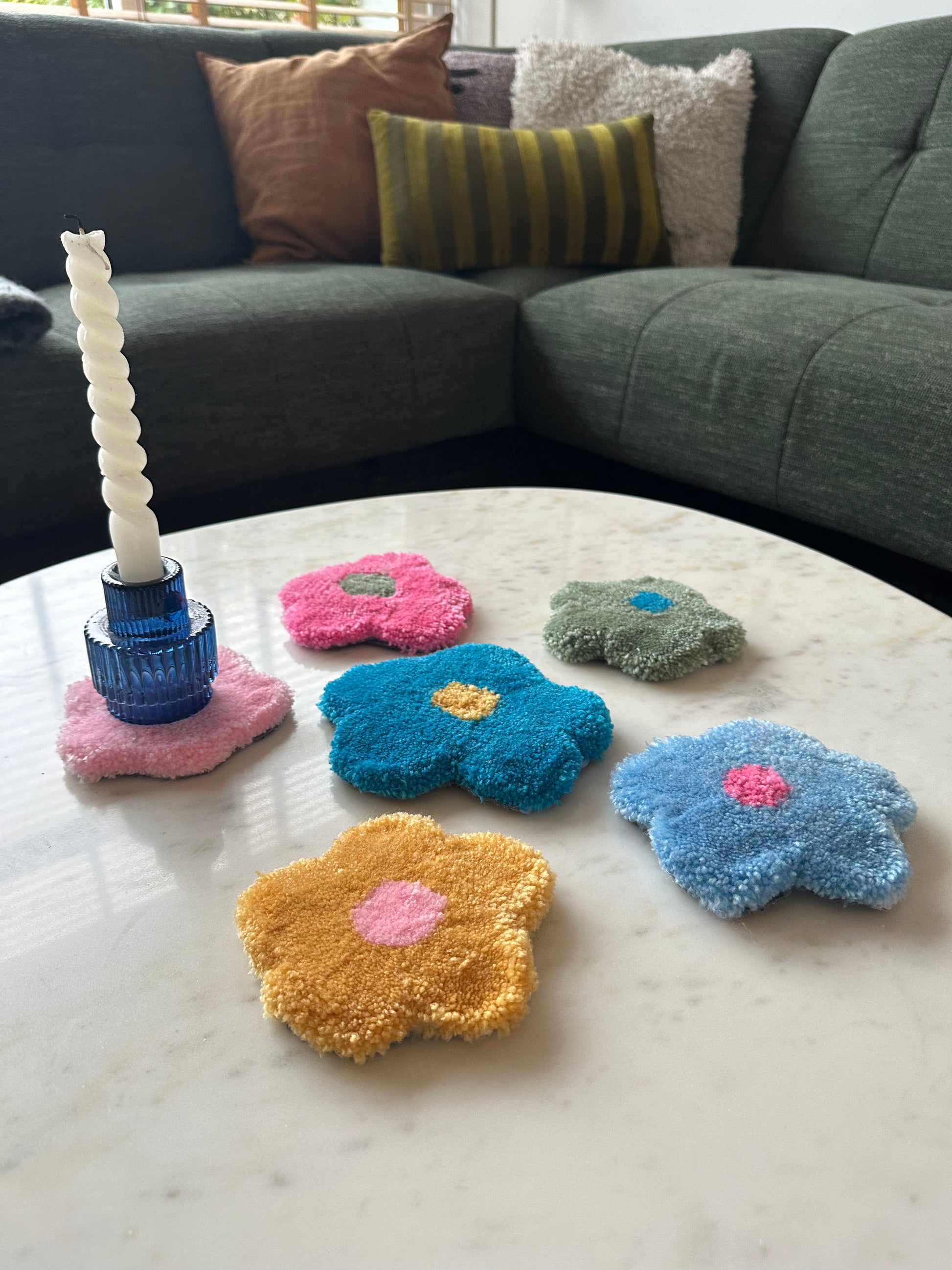 Our tufted flower shaped coasters placed on a table in various color combinations, such as green and blue, pink and green, yellow and pink, pink and blue, blue and pink. A blue candle holder with white candle is placed on one of the coastersMade by The Woollers