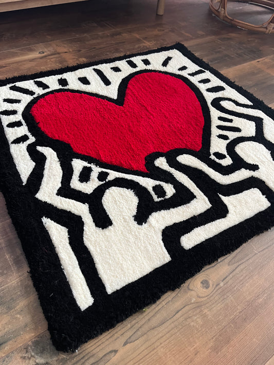 Shows our tufted rug of one of the artworks by Keith Haring. It is has black lining with white filling showing two people with a red heart in the middle. Made by The Woollers