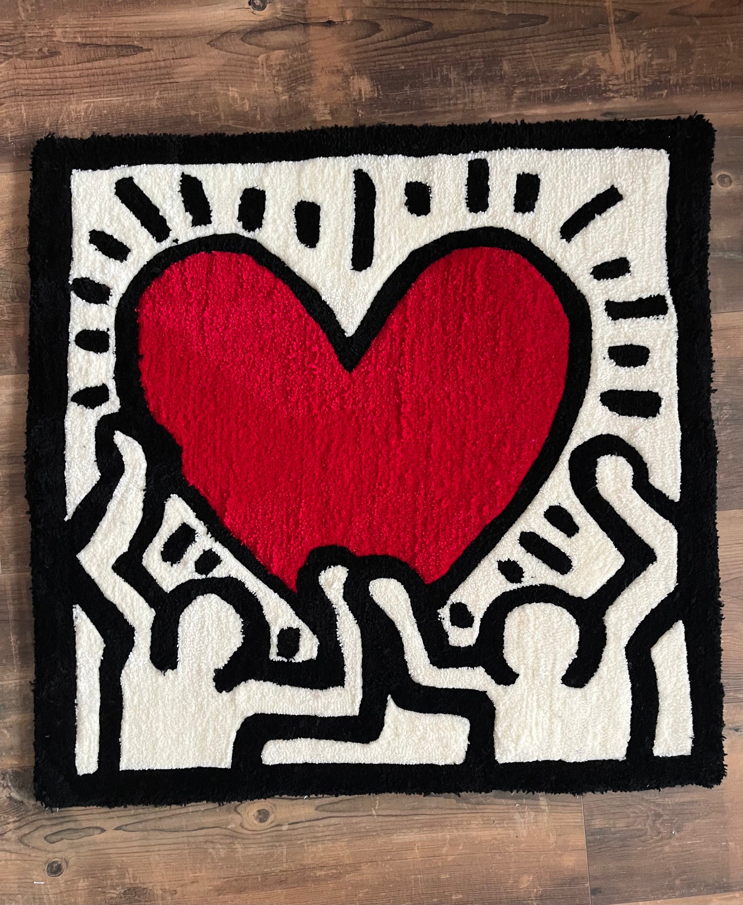 Shows our tufted rug of one of the artworks by Keith Haring. It is has black lining with white filling showing two people with a red heart in the middle. Made by The Woollers