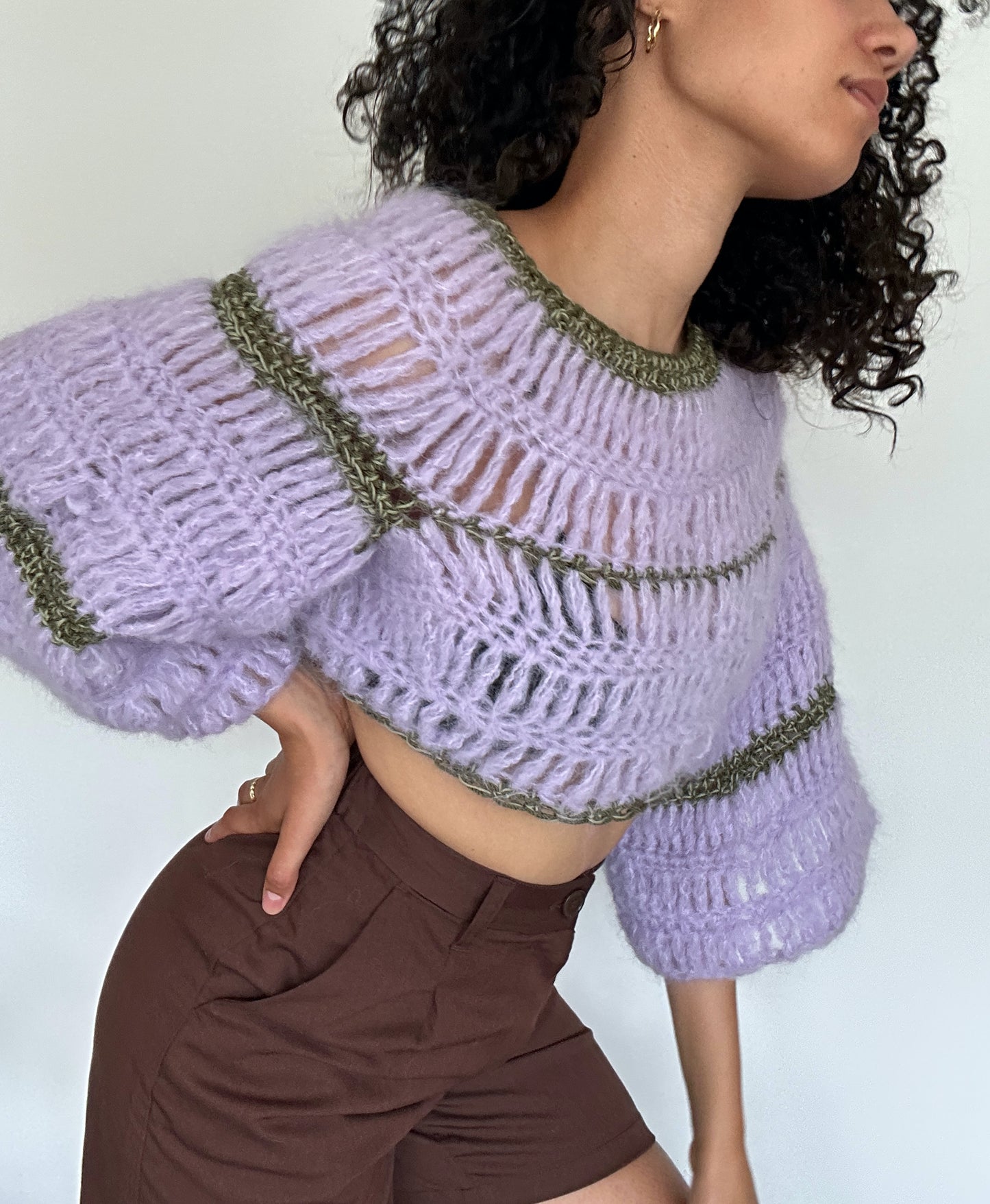 Shows a model wearing a lila and green coloured FLORA Long Sleeve Crop Top, which is crocheted and has a flowy design. Made by The Woollers