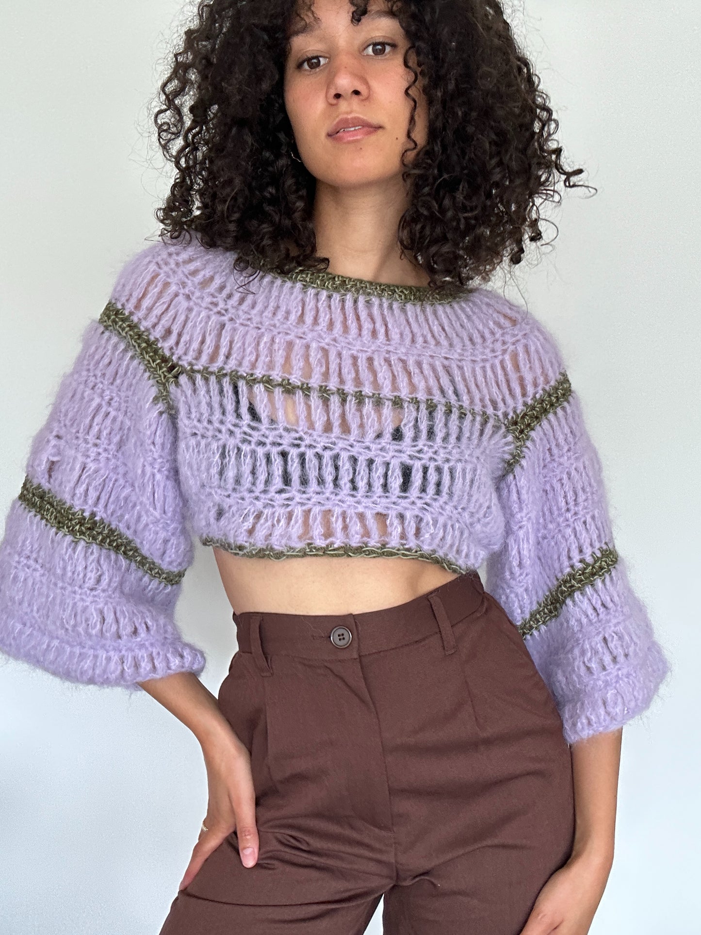 Shows a model wearing a lila and green coloured FLORA Long Sleeve Crop Top, which is crocheted and has a flowy design. Made by The Woollers