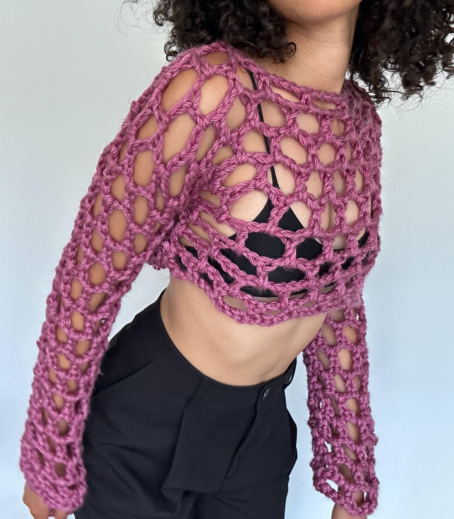 Shows a model wearing the crocheted KNOTTY Long Sleeve Crop Top, which is pink and has an open mesh design