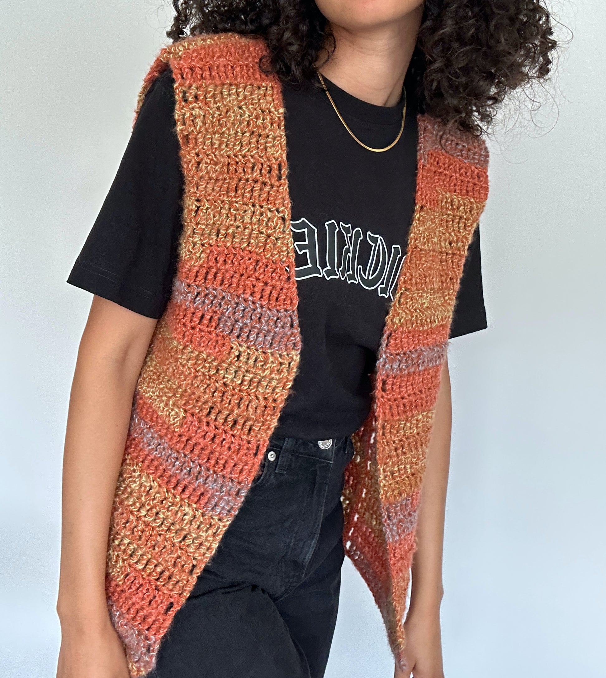 Shows a model wearing the crocheted MARI Open Vest which has hues of orange, yellow and blue. Made by The Woollers