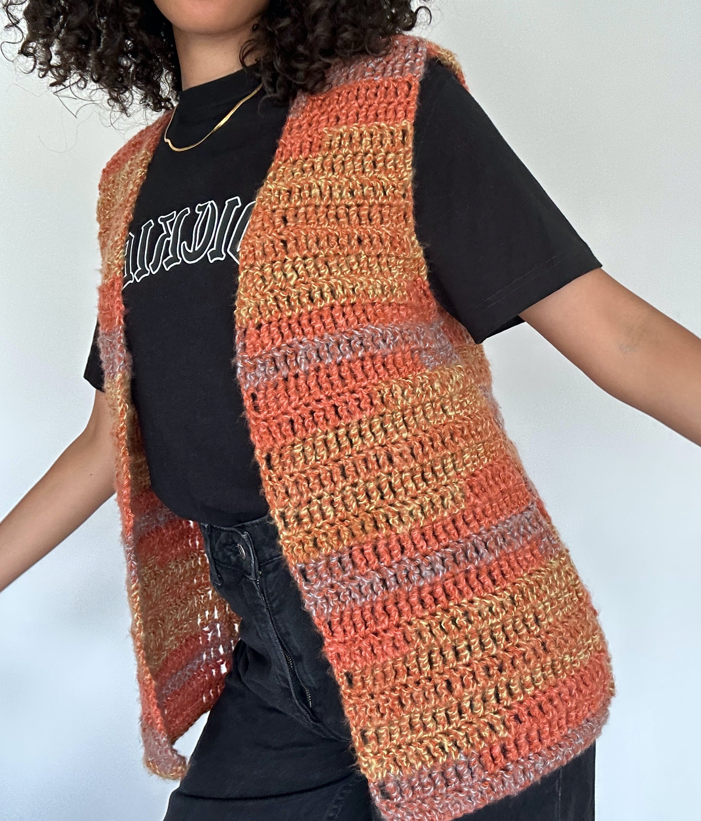 Shows a model wearing the crocheted MARI Open Vest which has hues of orange, yellow and blue. Made by The Woollers