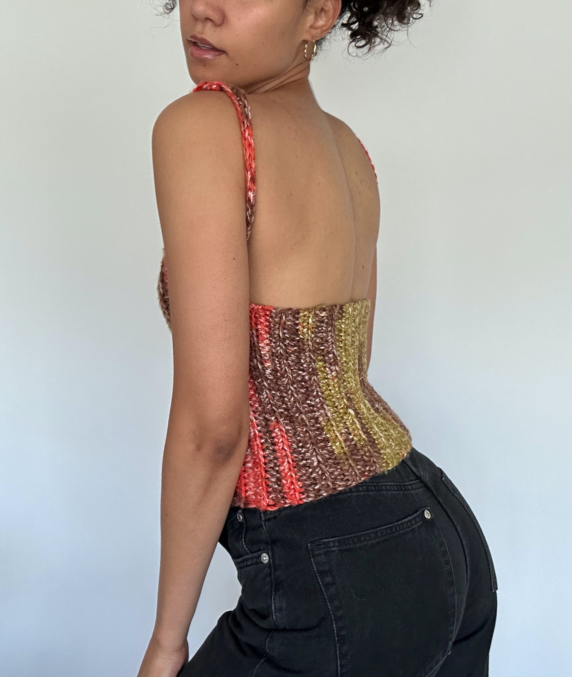 Shows a model wearing the crocheted NORA Open Back Top, which has a low cut open back and covered front in the colors brown, green and orange. Made by The Woollers