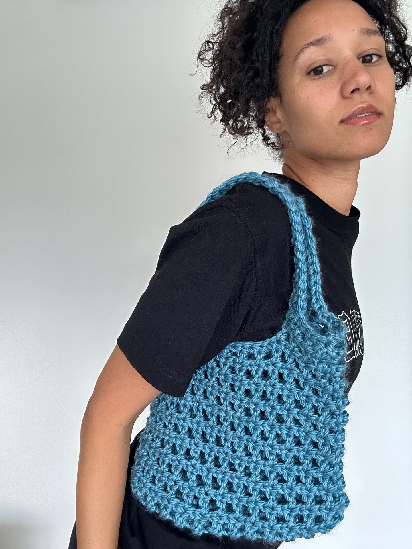 Shows a model wearing a medium sized blue crocheted KNOTTY Handbag which has an open mesh design. Made by The Woollers