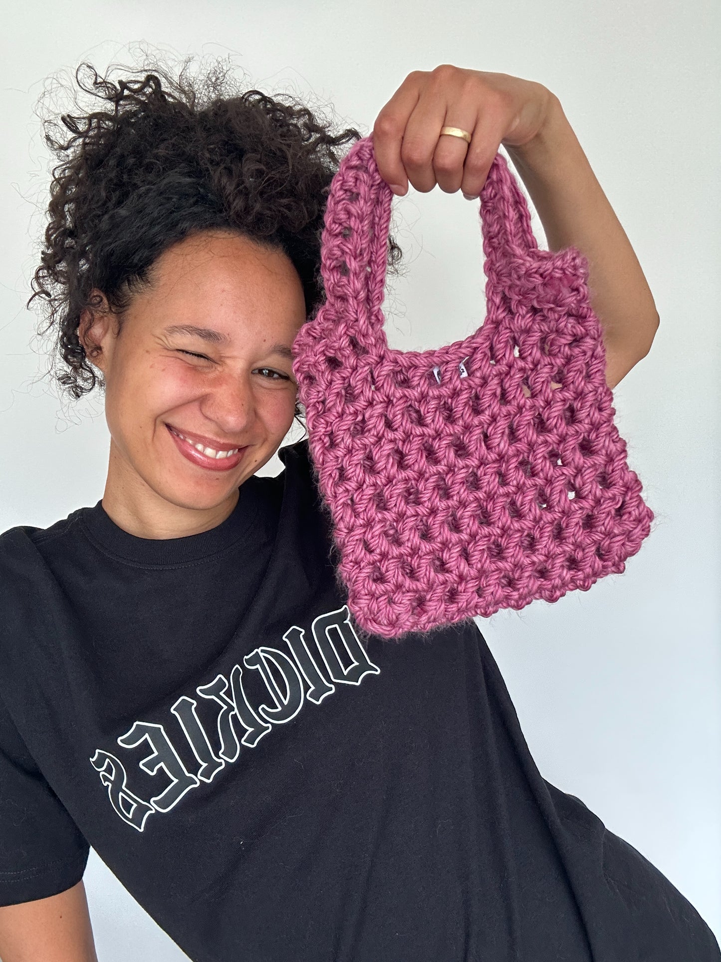 Shows a model holding a small pink crocheted KNOTTY Handbag which has an open mesh design. Made by The Woollers