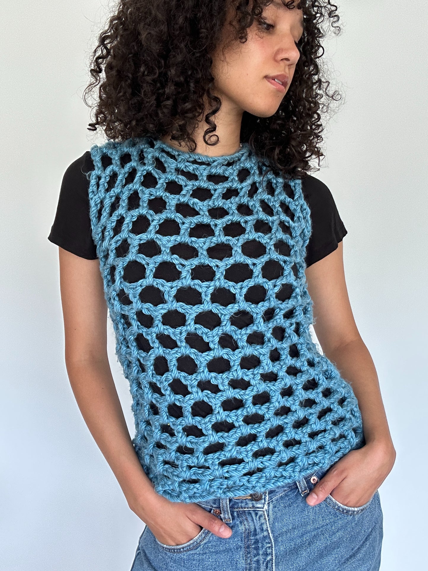 Shows a model wearing the KNOTTY Tank Top, which is a crocheted blue open mesh tank top made by The Woollers