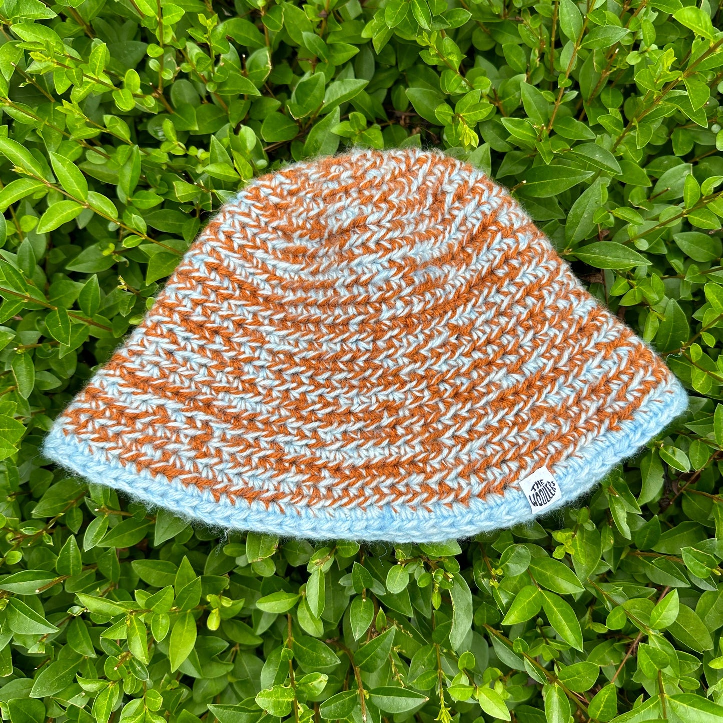 Shows a crocheted MOMI Buckethat in the colors blue and orange with a label that says 'The Woollers'