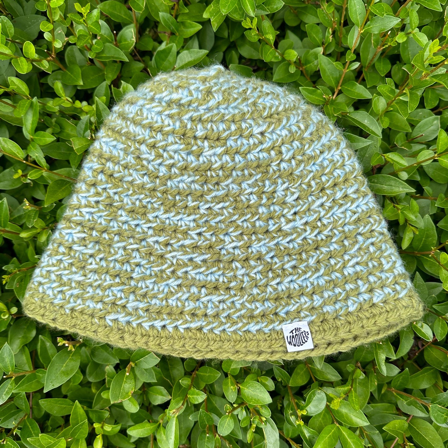 Shows a crocheted MOMI Buckethat in the colors green and blue with a label that says 'The Woollers'